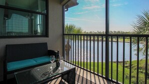 A balcony with a view of a lake, a glass table with a bottle and glasses, and a cushioned bench. - Exquisite 2 BR / 2 BA Lake Front Condo In Lakewood National Golf Club - Precious Gem (Lakewood Ranch)