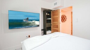 Un dormitorio con cama, televisor en la pared mostrando a un surfista, armario de madera y decoración geométrica en la pared.