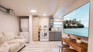 Una sala moderna con un sofá, una mesa de comedor de madera con taburetes, una cocina con electrodomésticos blancos y un televisor grande que muestra una escena tropical.