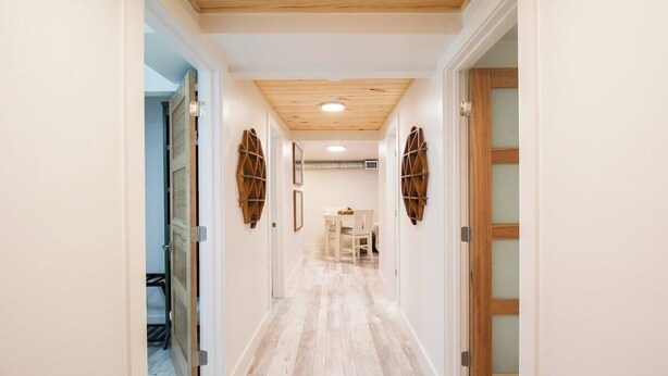 Un pasillo con piso de madera, paredes blancas y techo con vigas de madera. Hay dos puertas a cada lado, una con un panel de vidrio y la otra con marco de madera.