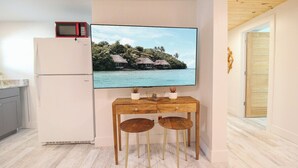 Cocina compacta con refrigerador blanco, microondas y una mesa de comedor de madera con dos taburetes. Un televisor de pantalla plana montado en la pared muestra una escena de una isla tropical.