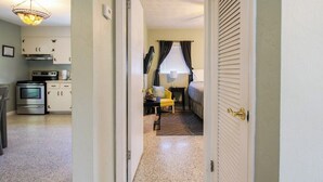 Un couloir mène à une chambre qui contient un lit, une chaise et une table de chevet. On aperçoit également un coin cuisine avec des placards et des appareils électroménagers à l’arrière-plan.