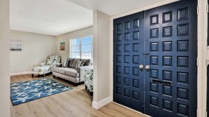Une pièce avec une porte bleu foncé, un canapé de couleur claire, un tapis à motifs et une fenêtre avec des stores.