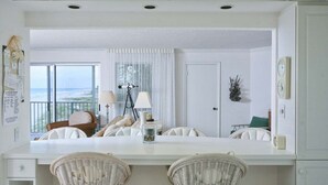 Een lichte, moderne kamer met een groot raam met uitzicht op de oceaan, een witte eettafel met rieten stoelen en een balkon met een telescoop.