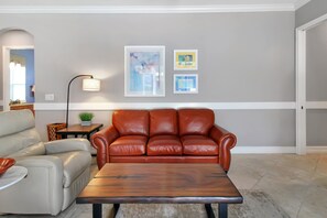 A living room with a brown leather sofa, a white armchair, and a wooden coffee table. There are framed artworks on the wall and a floor lamp. - Gorgeous lakefront pool home with golf course view Birdie (Lakewood Ranch)