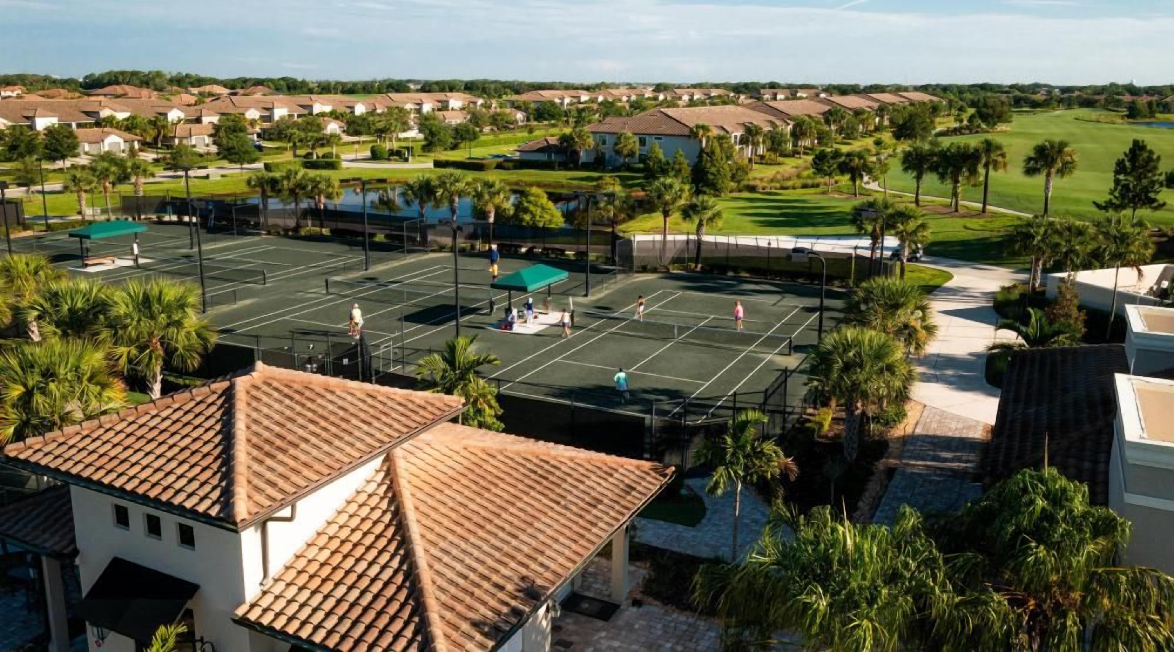 Uma quadra de tênis rodeada por edifícios residenciais, palmeiras e um campo de golfe ao fundo.
