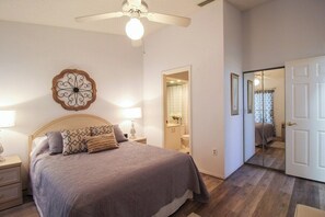 Una camera da letto con un letto, comodini, una ventola a soffitto, un elemento decorativo a parete, uno specchio in bagno e una porta che dà su un'altra stanza.