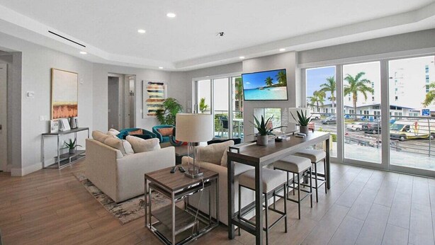 A modern living room with a sofa, coffee table, and a TV. There is a large window with a view of palm trees and buildings. - Luxury 4BR Siesta Key villa - Walk to the beach - Beach House Residences Unit 1 (Sarasota)