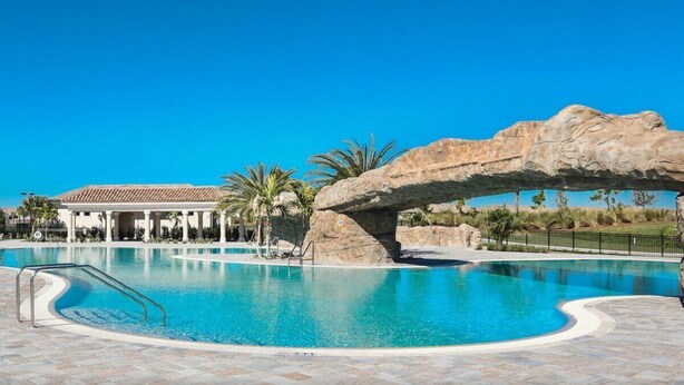 A swimming pool with a rock arch, palm trees, and a building with a tiled roof. - Lakefront 2 BR/2 BA condo in Lakewood National Golf Club - Pearl in the Rough (Bradenton)