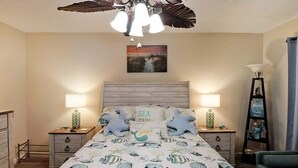 A bedroom with a bed featuring a sea-themed bedspread, two bedside lamps, a wooden headboard, and a ceiling fan.