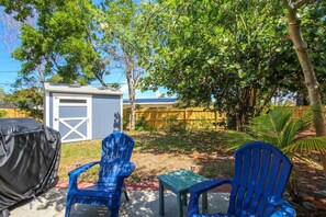 A backyard with two blue chairs, a small table, and a covered grill.