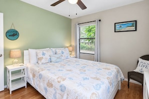 A bedroom with a bed, a nightstand, a chair, a window with blinds, a ceiling fan, and a wall with a framed picture.