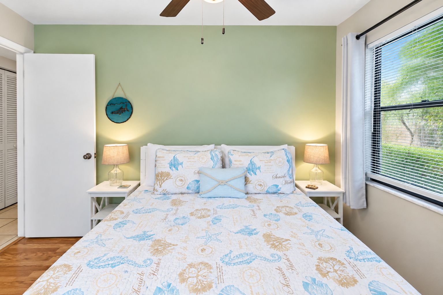 Une chambre à coucher comprenant un lit, deux lampes de chevet, un ventilateur de plafond, une fenêtre avec des stores et une porte.