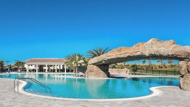 A swimming pool with a rock arch structure, palm trees, and a covered pavilion. - Breathtaking 3 BR condo with lake views in Lakewood National Golf Club - Shades Of Summer (Lakewood Ranch)