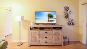 Een woonkamer met een flatscreen televisie op een houten kast, een vloerlamp en een decoratief kunstwerk aan de muur.