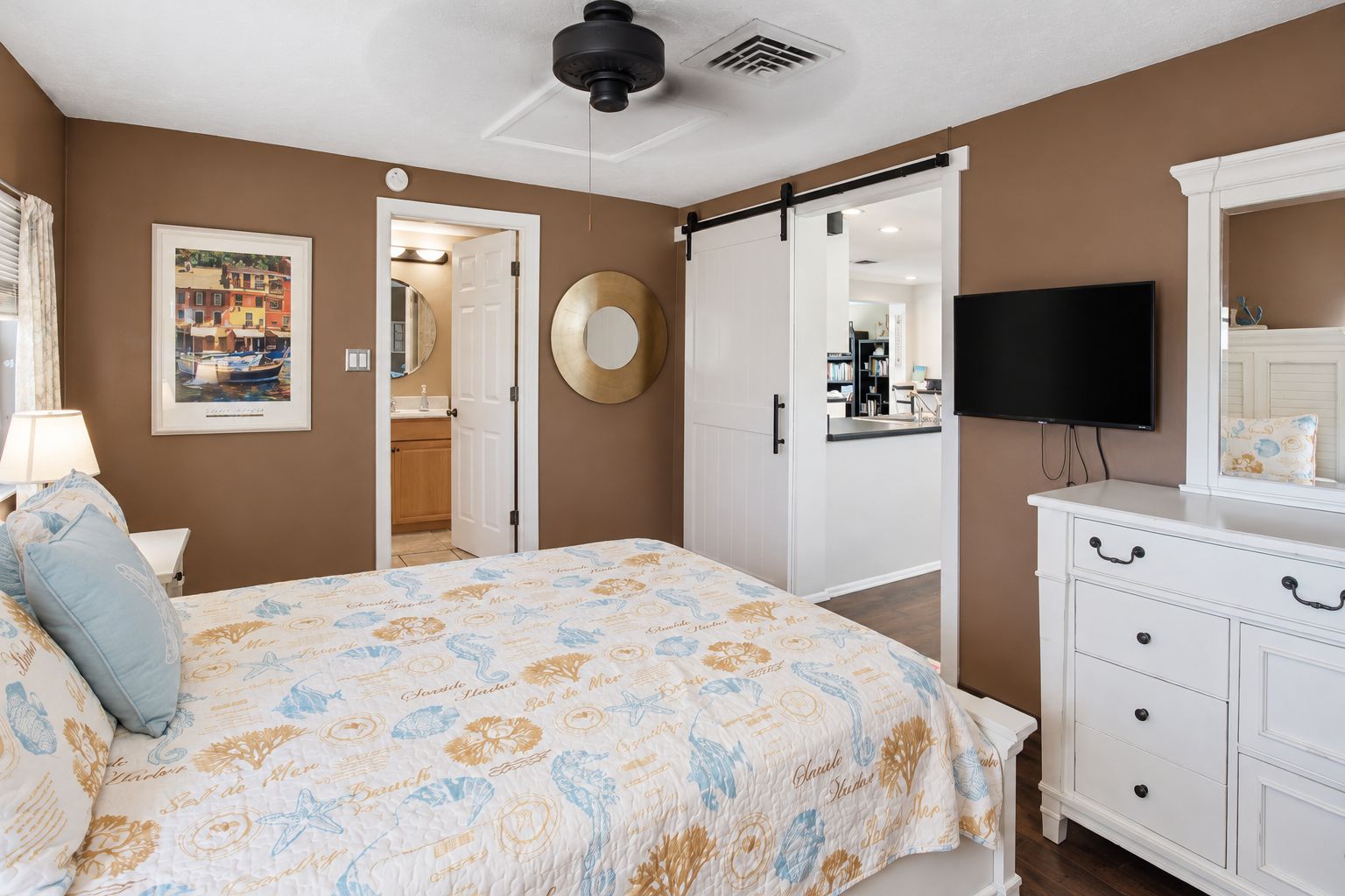 A bedroom with a bed, a dresser, a television, a door leading to a bathroom, and a wall-mounted mirror.