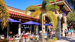 An outdoor cafe with tables and umbrellas, surrounded by palm trees and a building with a red-tiled roof. - 3 BR Pool Home In Nokomis - Close To Beaches - Tropical Escape (Nokomis)