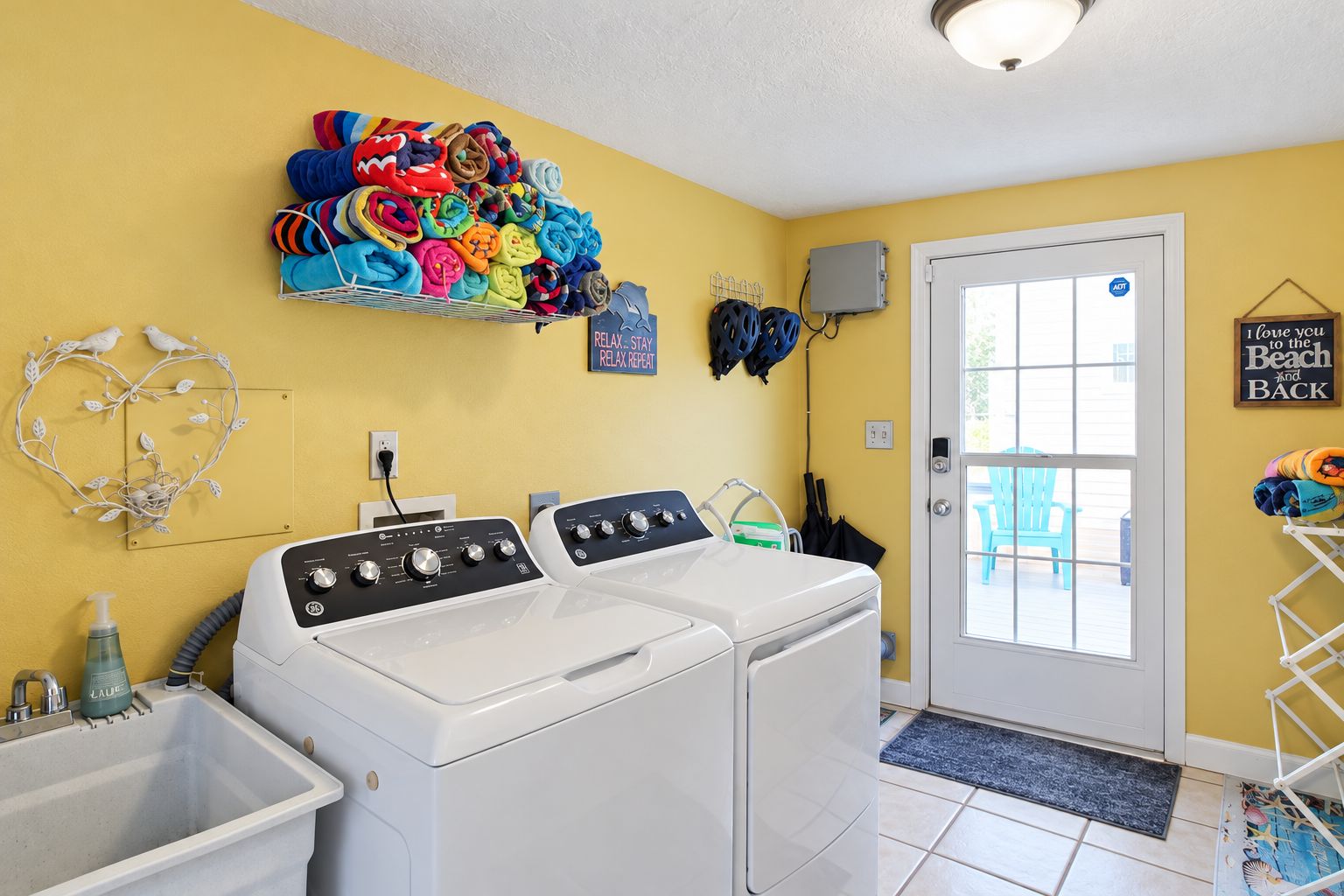 A laundry room with two front-loading washers, a drying rack with colorful towels, a heart-shaped decoration, and a door with a view to the outside.