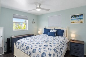 Une chambre à coucher comprenant un lit, deux tables de chevet, un ventilateur de plafond, une fenêtre avec des stores et un luminaire fixé au mur.