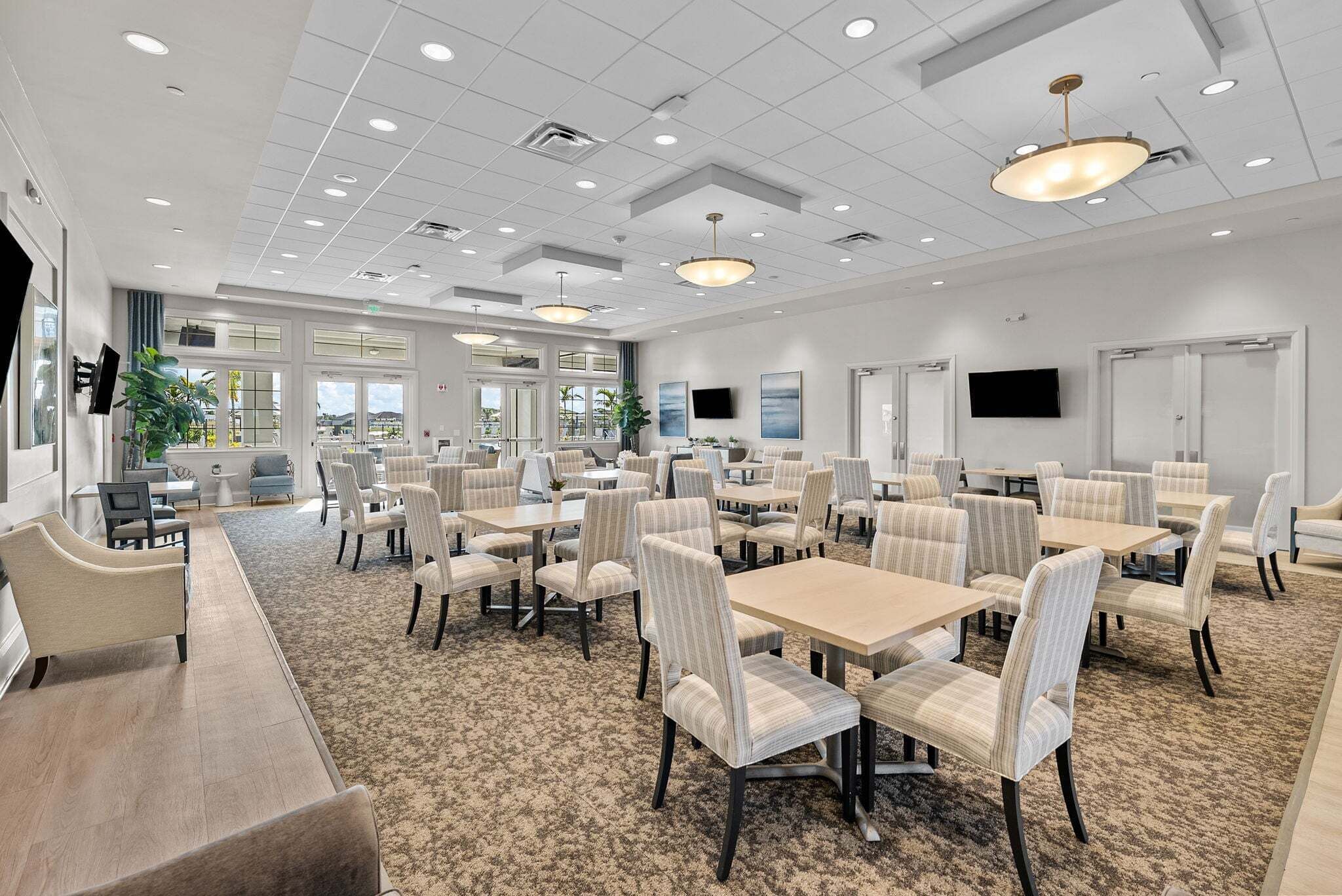 A spacious hotel dining area with multiple tables and chairs, ceiling lights, and a large window providing natural light.