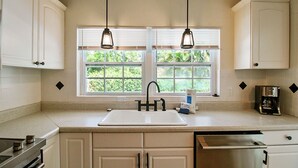Una cocina con armarios blancos, un lavamanos doble, una cafetera y una ventana con persianas.