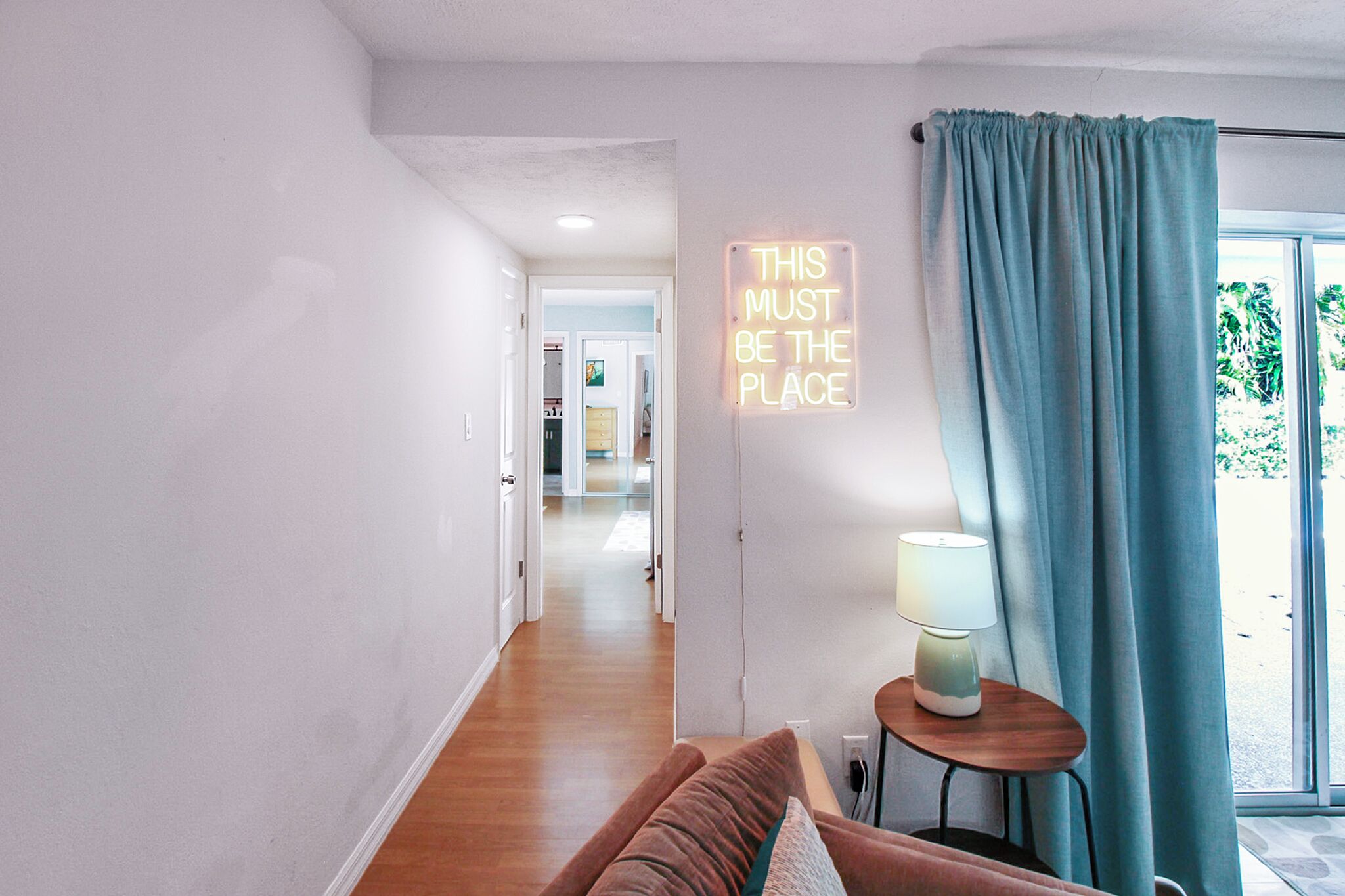 Un interno moderno con un corridoio che conduce a una stanza, un neon al muro, una tenda blu e una lampada su un tavolino.