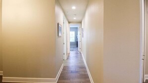 A narrow, well-lit hallway with beige walls and a wooden floor, leading to a door at the end.