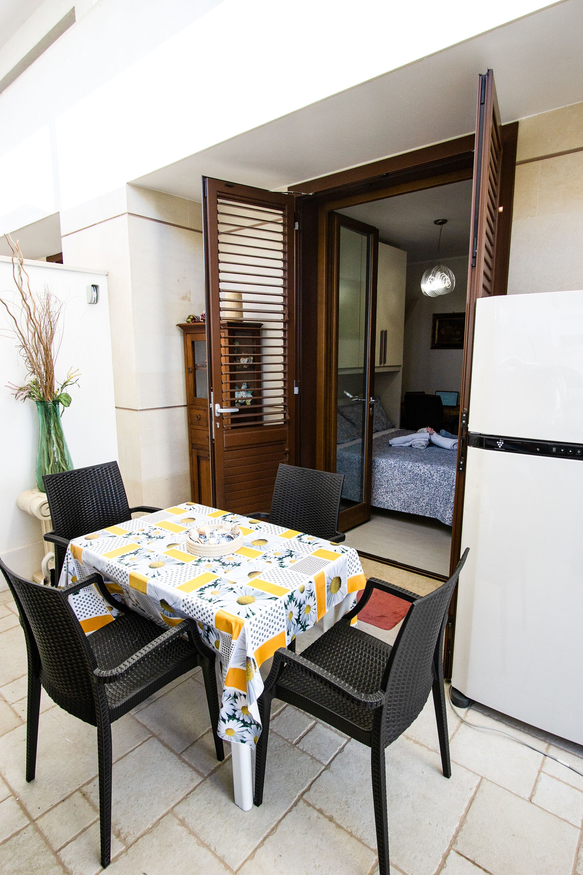 A small dining area with a table and four chairs, a refrigerator, and a bed visible through an open door.