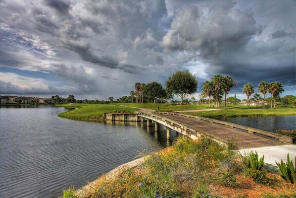 A serene lakeside view with a wooden bridge, lush greenery, and a cloudy sky. - Golf course view - 2BR - 2nd floor condo - Eagle Hunters (Sarasota)