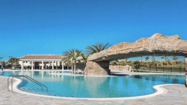 A swimming pool with a rock arch, palm trees, and a covered area in the background. - Exclusive 2 BR Lakefront Condo in Lakewood National - The Duffers Retreat (Lakewood Ranch)