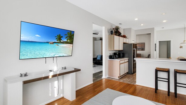 A modern kitchen with a large island, a dining area with a white table and bar stools, and a TV displaying a beach scene. - Luxurious 4BR 2BA home with pool Bradenton My Great Escape (Bradenton)