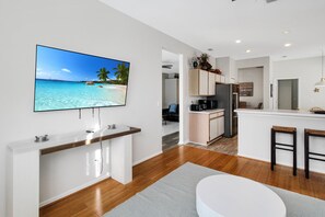 Een moderne keuken met een groot eiland, een eethoek met een witte tafel en barstoelen, en een televisie waarop een strandscène wordt weergegeven.