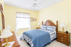 Une chambre à coucher comprenant un lit, deux tables de chevet, un ventilateur de plafond, une fenêtre avec des rideaux et un miroir.