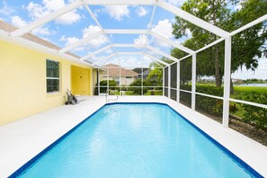 Une piscine dotée d’une terrasse blanche, entourée d’une structure en verre, et offrant une vue sur un quartier résidentiel avec ses maisons et ses arbres.