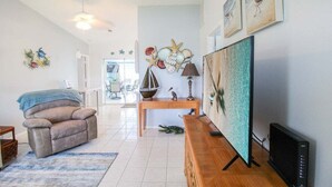 Ein Wohnzimmer mit Liegekorbstuhl, Couchtisch aus Holz, Fernseher und Wandbildern mit Möwen und Muscheln.