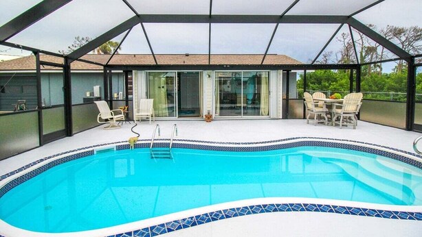 Ein überdachter Swimmingpool mit gefliestem Rand, umgeben von einer Glaskonstruktion, mit Gartenstühlen und einem Tisch ausgestattet.