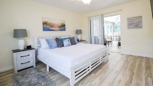 A bedroom with a white bed, a nightstand, a lamp, a painting, and a view of a dining area through a glass door. - Gorgeous 2BR 2BA Private Pool Home Near Siesta Key Sweet Pineapple (Sarasota)