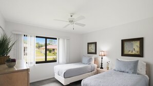 A bedroom with two beds, a ceiling fan, a window with blinds, a bedside table with a lamp, and a framed picture on the wall.