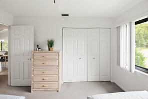 A bedroom with a bed, a dresser, a closet, and a window with a view of greenery.