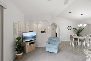 A modern living room with a flat-screen TV, a light blue armchair, and a dining area with white chairs and a table.