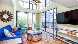 A spacious living room with a blue sofa, wicker ottomans, a large TV, and a balcony with outdoor furniture. - Spacious 3BR Retreat On Sanibel Island - Steps to the beach - Cyprina Beach #10 (Sanibel)