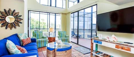 A spacious living room with a blue sofa, wicker ottomans, a large TV, and a balcony with outdoor furniture.