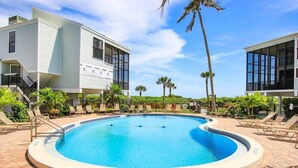 A swimming pool surrounded by lounge chairs and palm trees, with a modern building in the background. - Spacious 3BR Retreat On Sanibel Island - Steps to the beach - Cyprina Beach #10 (Sanibel)