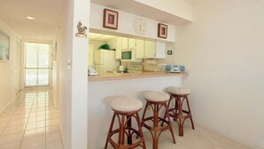 Een keuken met witte kastjes, een magnetron en een klok aan de muur. Er staan drie houten krukken met beklede zitjes.