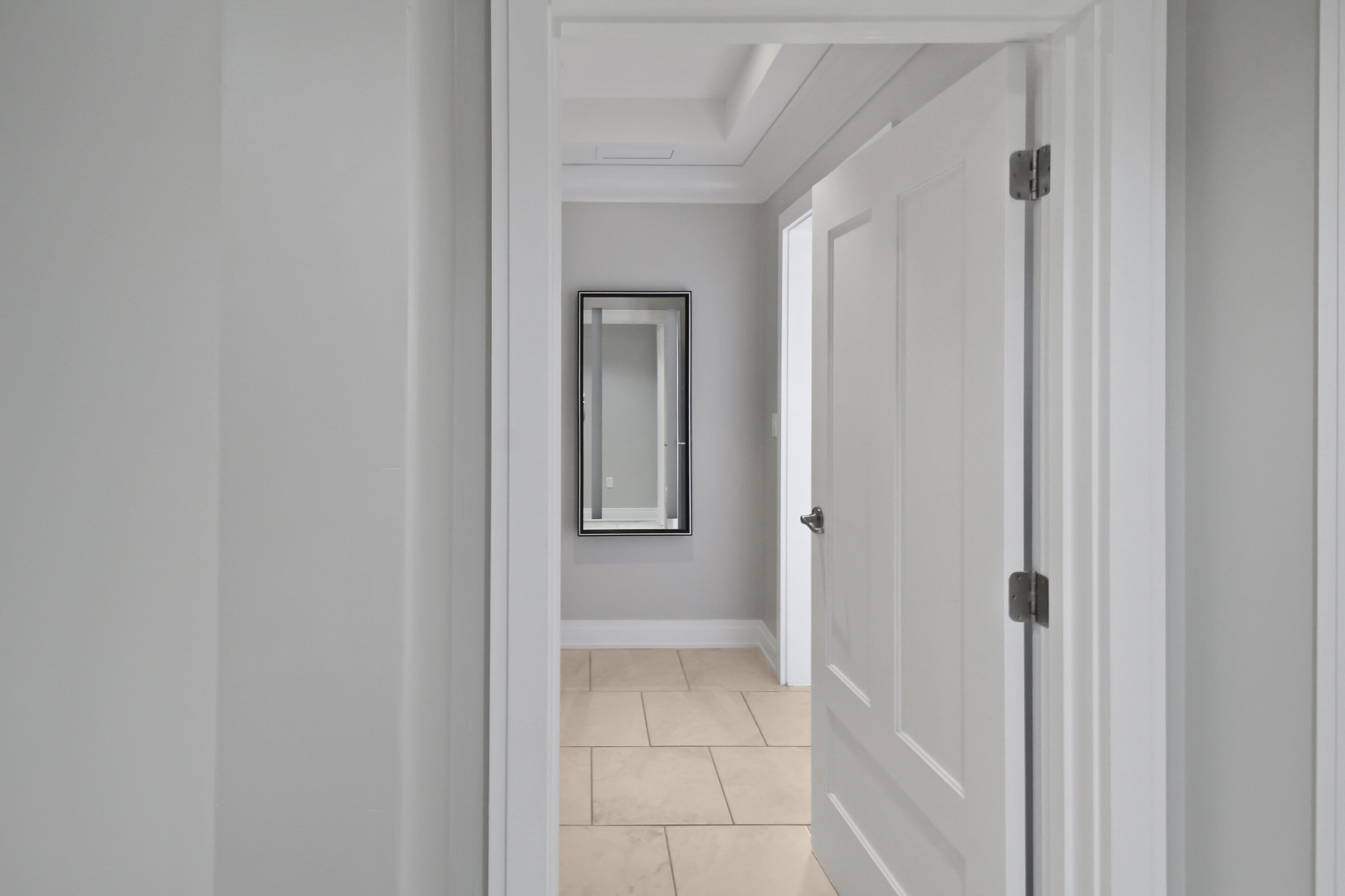 A hallway with a door on the right, a framed mirror on the wall, and a tiled floor.