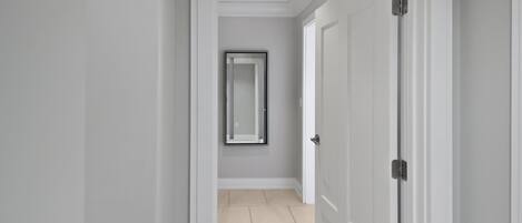 A hallway with a door on the right, a framed mirror on the wall, and a tiled floor.