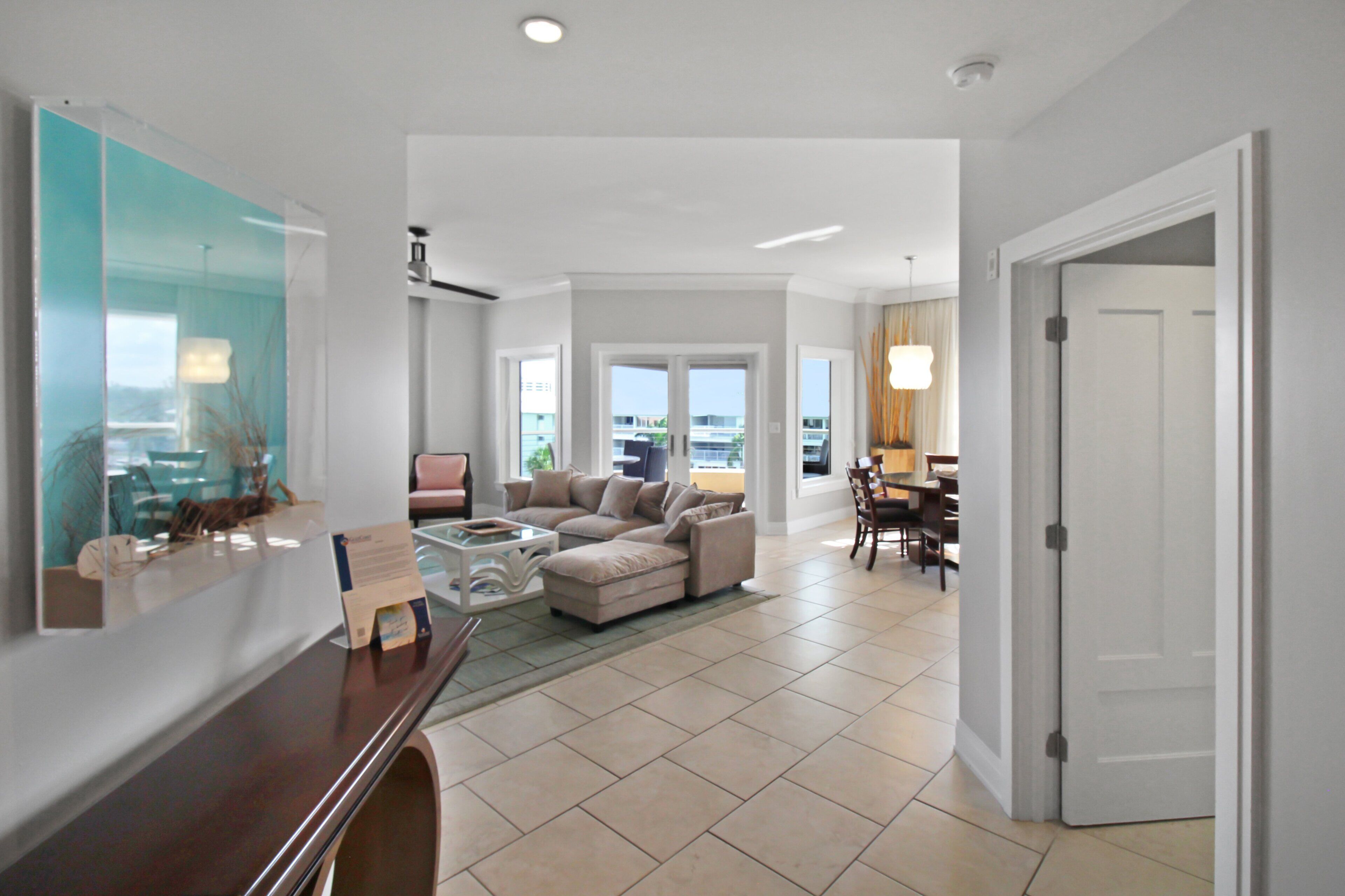 A spacious living area with a large sofa, a coffee table, and a dining table with chairs. There is a door leading to another room and a view of the outside through a window.