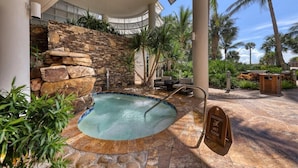 Zona de piscina con muro de piedra, palmeras y un letrero de precaución sobre el piso mojado.