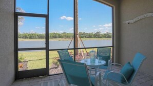 Habitación con un ventanal grande que ofrece vistas a un lago y vegetación. Hay una mesa redonda con sillas y una mesita con un florero y vasos.