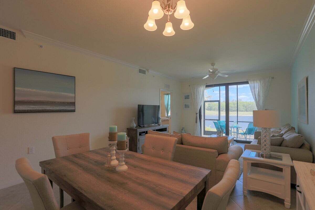 Una sala de estar con una mesa de comedor de madera, sofás color beige, un televisor y un ventanal con cortinas.